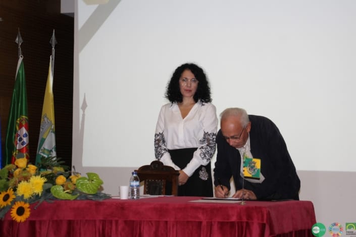 Homem de óculos e fato escuro assina documento em mesa com bandeiras; mulher de blusa branca observa.