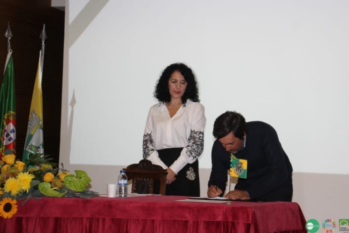 Duas pessoas em cerimónia oficial junto a mesa com bandeiras e flores amarelas.