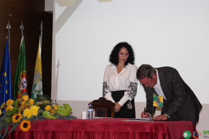 Homem de fato assinando documento numa cerimónia oficial, com mulher ao lado e bandeiras ao fundo.