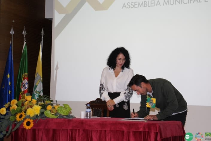 Dois participantes em cerimónia oficial na Assembleia Municipal, com bandeiras e flores amarelas na mesa.