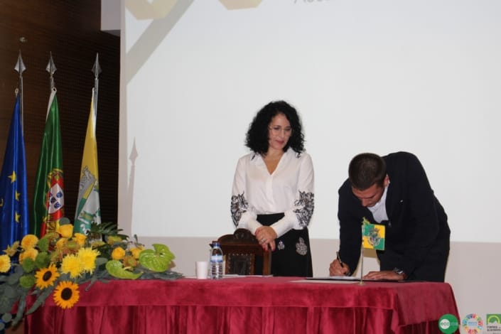 Homem de fato preto assina documento em mesa com bandeiras e flores amarelas.