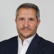 Homem de meia-idade, cabelos grisalhos, barba, camisa branca e blazer azul-marinho.