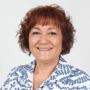 Mulher de meia-idade com cabelo castanho encaracolado, sorriso aberto, veste padrão azul e branco, fundo cinzento neutro.