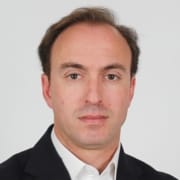 Homem de meia-idade, cabelo castanho, com ligeira calvície frontal, veste fato preto e camisa branca, fundo neutro cinzento.