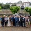 Grupo de pessoas em formação para foto de homenagem num pátio com muro de pedra e edifício ao fundo.