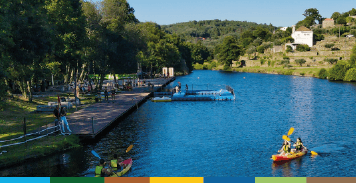 Praia fluvial com cais de madeira, embarcação amarela ao fundo e zona arbórea envolvente.