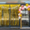 Entrada de estabelecimento decorada com cortina de franja dourada e balões rosa, branco e amarelo em guirlanda